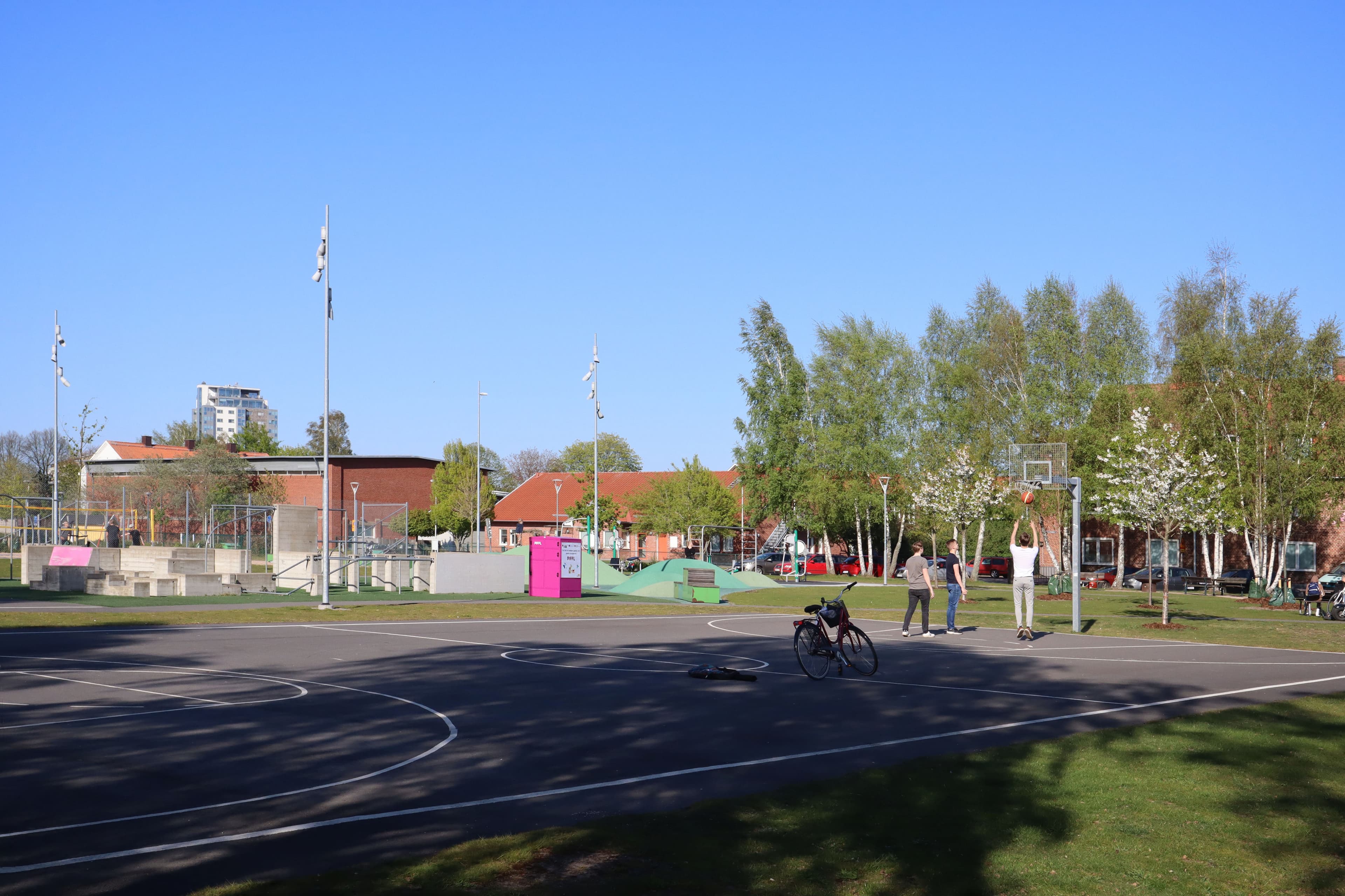 Spontanbasket i parken – Hämta din boll från en Pifflbox Tre killar spelar basket vid en Pifflbox i en park under en solig dag